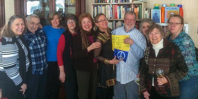 A group of middle-aged and senior people standing in front of a school room bookcase, holding out a book with a peace symbol on the cover
