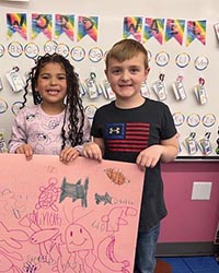 2 young children hold up their poster. A classroom whiteboard is behind them with colorful ribbons and smiley faces.