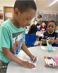 A school-aged boy in a teal t-shirt draws a poster at a classroom art table.