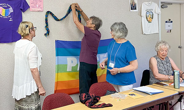 An older woman pins a hand knitted scarf to a classroom pin board. There is a pride flag and t-shirt pinned on the board, as well. Several other women look on.