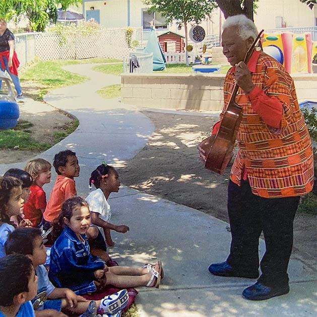 An older woman in an orange patterned top plays guitar for young children seated on the ground in front of her.