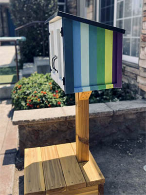 A side view of a little free library — a covered box, approximately 2 feet wide and 2 feet tall and 1 foot deep. It sits on a mailbox post on a wooden platform outside. The side is painted with stripes of purple, blues, and greens.