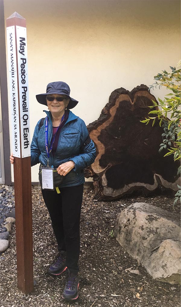 Irene Lipshin wearing outdoor weather gear stands next to a pole in a garden. The pole reads, "May Peace Prevail on Earth."