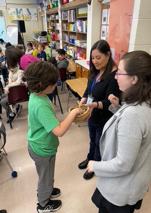 Kim Moberg and Heather Swanson offer a basket with treats in it to a child in a green t-shirt in the classroom.