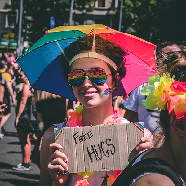 Person in an outdoor parade wearing rainbow glasses, a rainbow umbrella hat, and with a rainbow sticker on their check is holding a cardboad sign that says "Free Hugs"