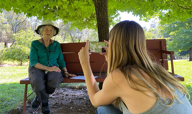 A college-aged woman is in the foreground, kneeling down to record an older woman with her phone. The woman is seated on a park bench in the shade.