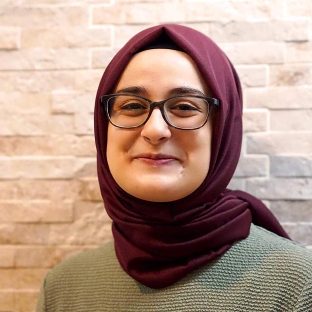 A 30-year-old woman wearing a burgundy hijab and glasses smiles for the camera