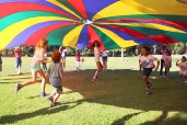 Children running under a rainbow-colored tarp on a grassy field. Other children are holding the tarp at the edges and waving it up high, then down to the ground.