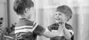 Two toddler boys hold hands and look at each other while laughing.