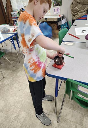 Young boy stands at a classroom table making a Tibetan bowl sing.