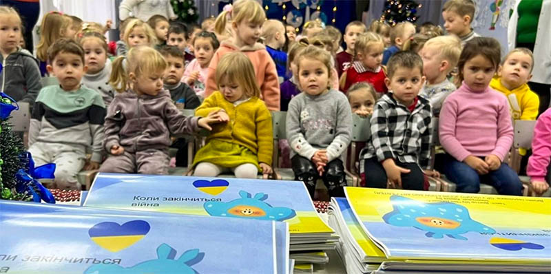 Preschool-aged children sit in chairs waiting to be handed the books on the table in the foreground.