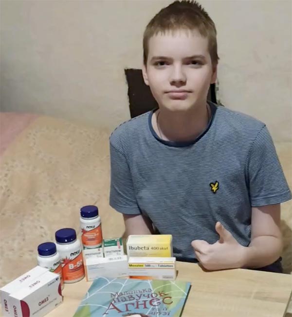 A young boy sits at a table giving a thumbs up sign to the viewer. On the table are boxes and bottles of medicines and a children's book.