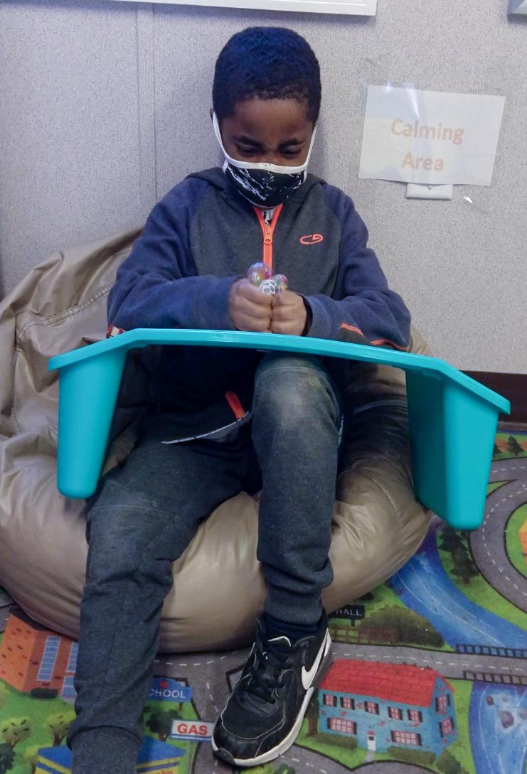 A black school-aged boy with a mask on sits in the calmness area using a plastic lap tray as a drawing surface