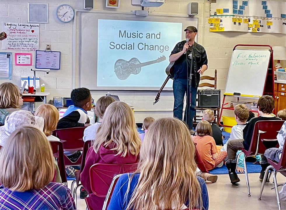 A middle-aged man in jeans long-sleeved-shirt and a baseball cap stands in front of a room of school-aged children. He has a guitar slung over his right shoulder and a slide shines on the wall behind him reading "Music and Social Change"
