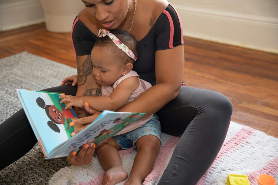 A mother reading to her daughter. They are seated on a blanket on the floor.