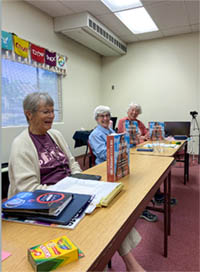 A group of older women seated at a long school desk explaining the children's books in front of them on the desk.