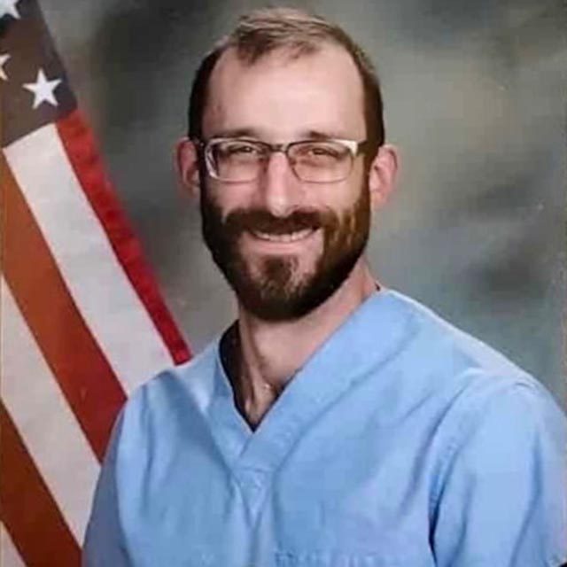 Alex Preti, in his blue scrubs, in front of a US flag.