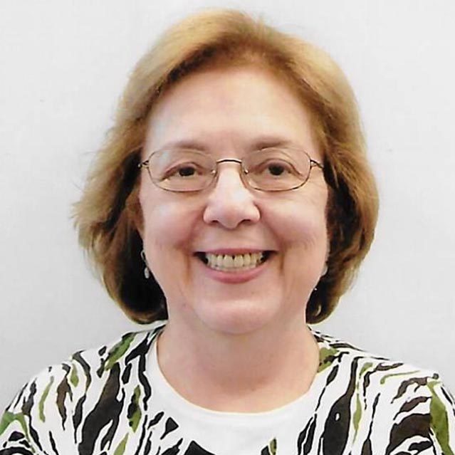 Headshot of an older, Caucasian woman with auburn chin length hair. She wears glasses and a green jungle-print blouse.
