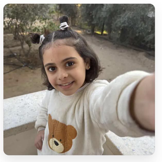 A young Palestinian girl wearing a teddy bear sweatshirt holds takes a selfie.