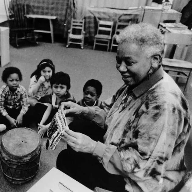 Black and white photo of Ella Jenkins seated in a classroom with young children seated on the floor watching her. There is a drum at her knees.