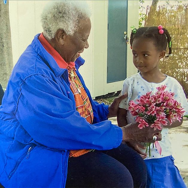 An older woman in a bright blue shirt receives a bouquet from a young girl in pigtails.