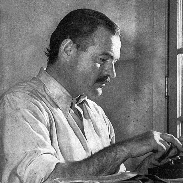 Black-and-white portrait of Ernest Hemingway from the side as he types a manuscript.