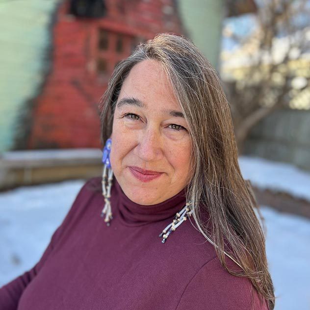 Headshot of a middle-aged woman with long greying hair wearing a burgundy turtleneck and long earrings.