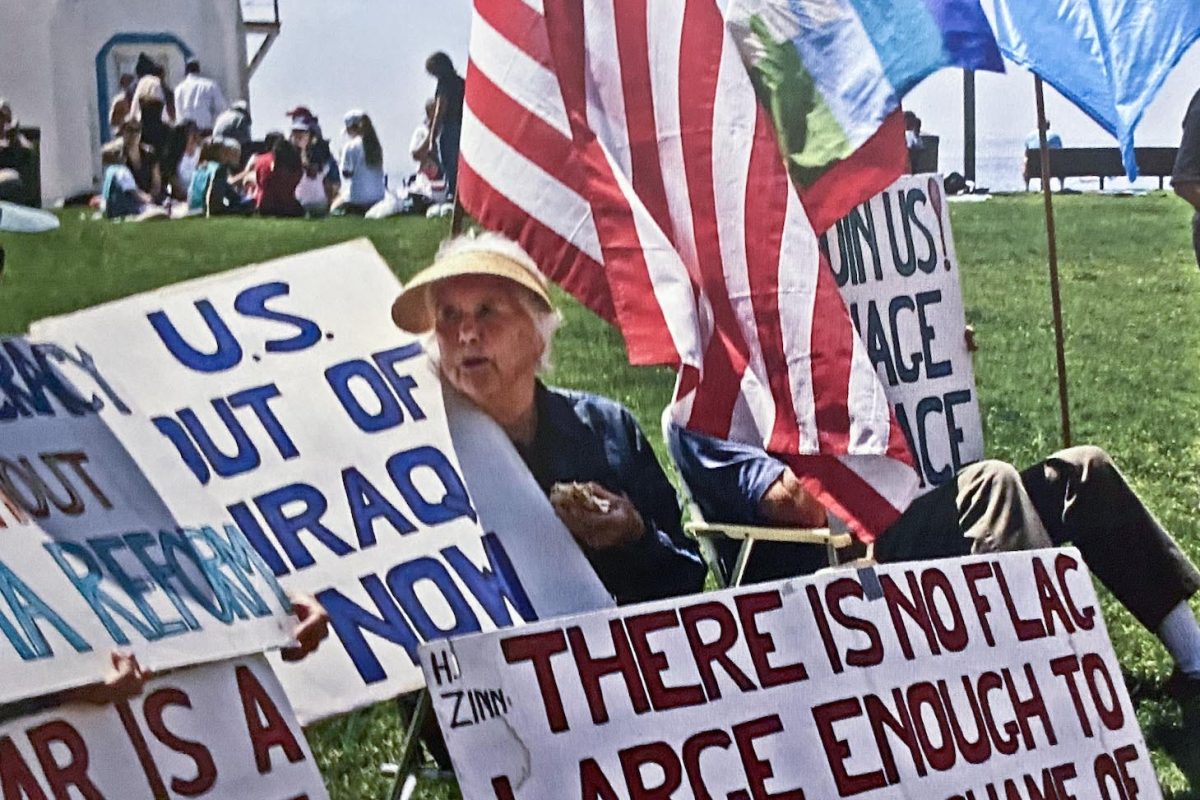 An older woman is seated outside at Laguna's Main Beach holding a US flag. Protest signs lean across her legs and other chairs.