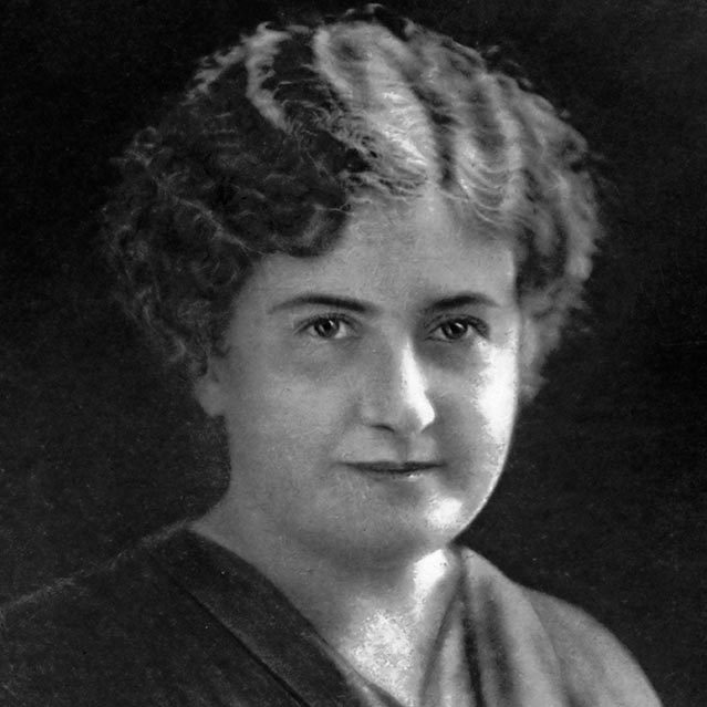 An antique black-and-white photo of a middle-aged woman with short hair wearing a v-necked dress.