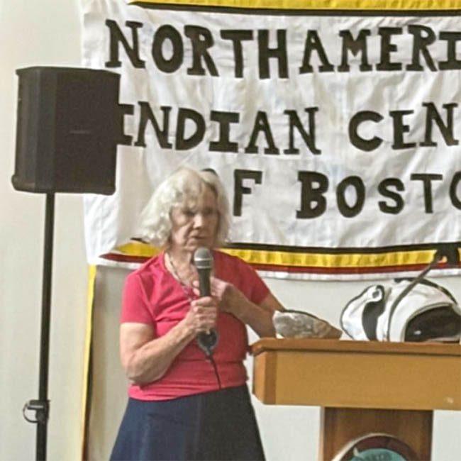 Pat Landry, an older Caucasian woman with chin-length white hair, stands at a podium holding a microphone. A handmade banner behind her reads 