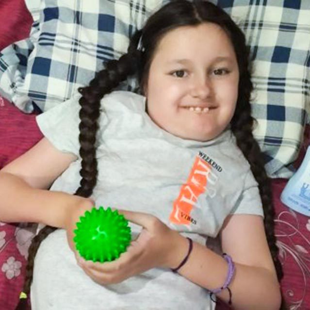 A Ukrainian orphan leans back against a plaid pillow, smiling up at the camera. She has breaks and holds a green spiked dryer ball.
