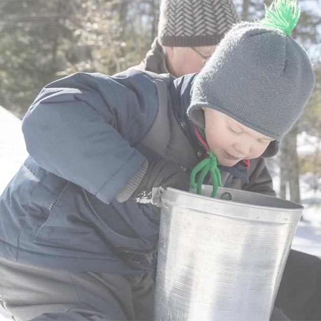 Screenshot showing a young boy outside in a snowsuit looking down into a silver bucket.