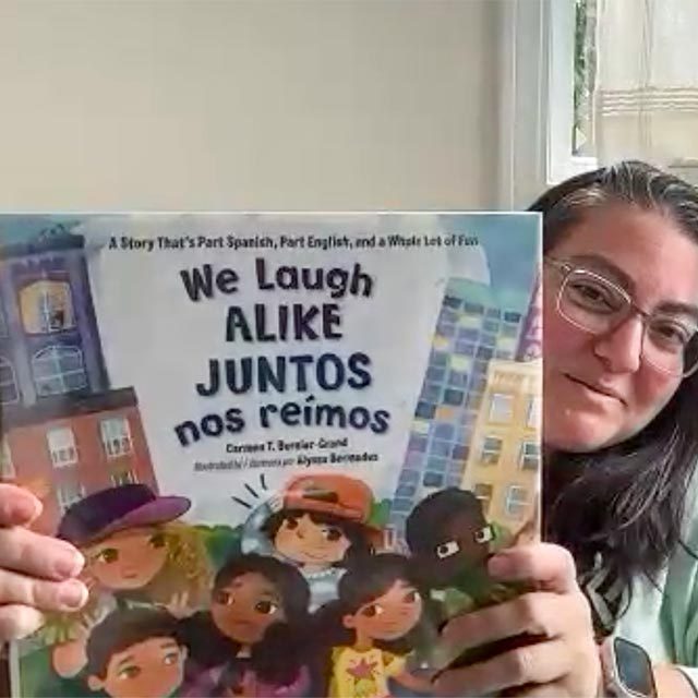 Screen capture of Monica holding the book We Laugh Alike, Juntos nos reimos, up to the computer camera.