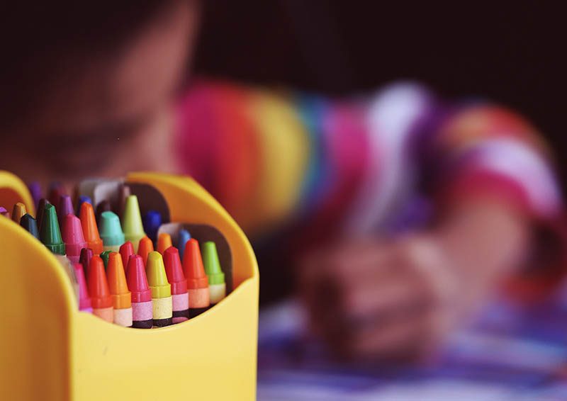 Close up on the tops of crayons in a plastic yellow box. Soft focus in the background a child in a brightly colored long-sleeved striped shirt draws with a crayon