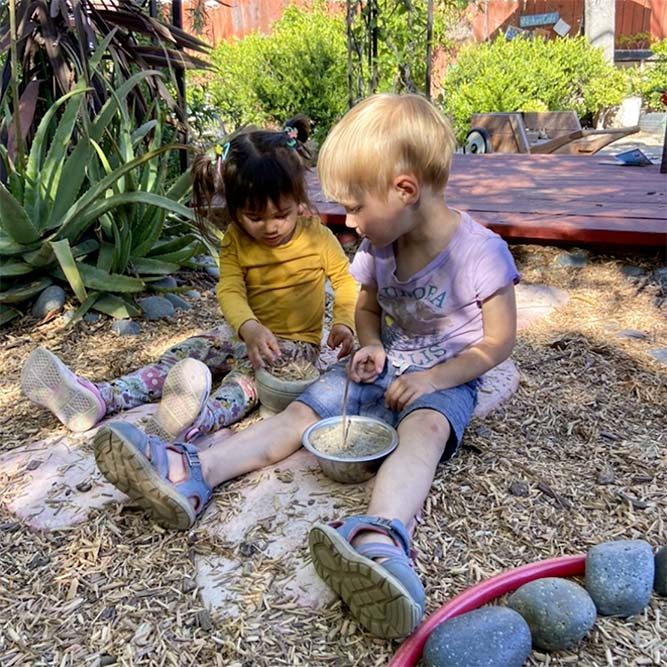 2 toddlers seated outside on the ground playing with loose parts in bowls