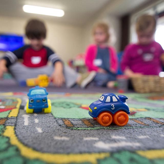Close up of 2 toy cars on a rug depicting roads. Three young children are soft focus in the distance.