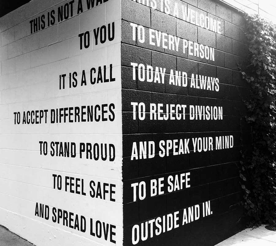 Two sides of a brick wall, seen from the corner. One side is painted white with black text, the other is painted black with white text. The two walls read: This is not a wall, this is a welcome to you to every person. It is a call today and always to accept differences to reject divisions, to stand proud and speak your mind. To feel safe, to be safe.