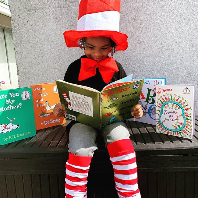 A child dressed as Cat in the Hat sitting on a bench reading Dr. Seuss books.