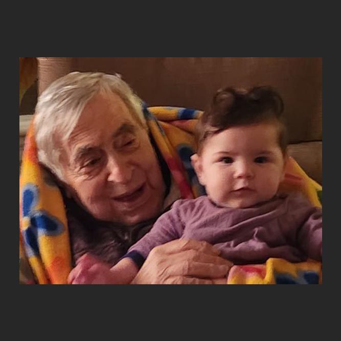 A senior man smiles at a toddler in his arms. There's a colorful floral blanket around their shoulders.