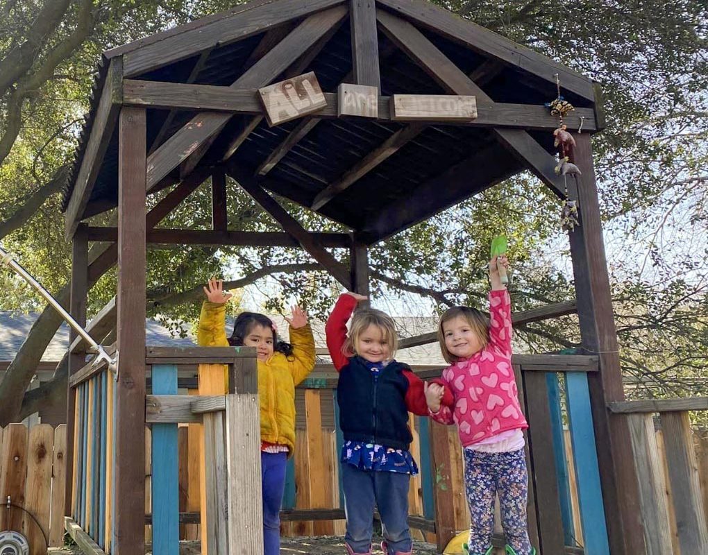 3 girls standing in their open wood clubhouse. All 3 are smiling and pointing upward to the sign that reads 