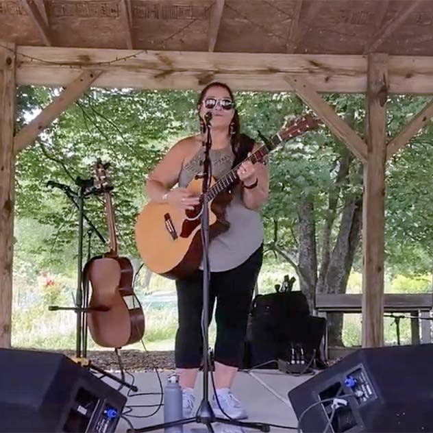 A young woman standing on an outdoor stage playing an acoustic guitar and singing into a microphone.