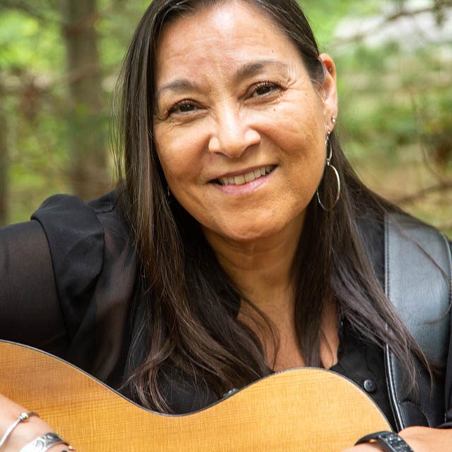 A middle-aged woman with long, straight black hair holding a guitar.