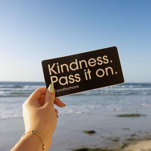 A hand holding up a small sign that says Kindness. Pass it on. The shoreline is visible in the distance.