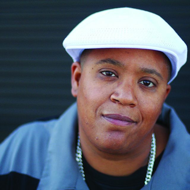 headshot of a black person with a white beret, a grey collared jacket, and a thick silver chain around their neck.