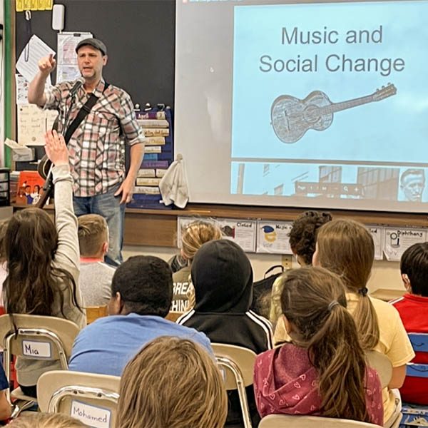A man with a guitar stands in front of a class of elementary school children speaking into a mic, discussing the slide 