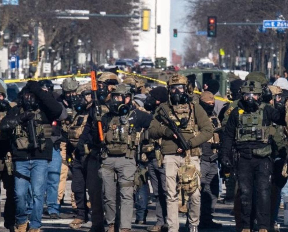 ICE officers and vehicles stand blocking a large intersection. They wear gas masks and hold weapons and other military gear.