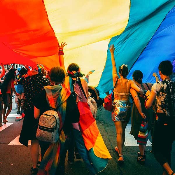 A group of colorfully dressed people dance under an enormous flag as they parade down the street.