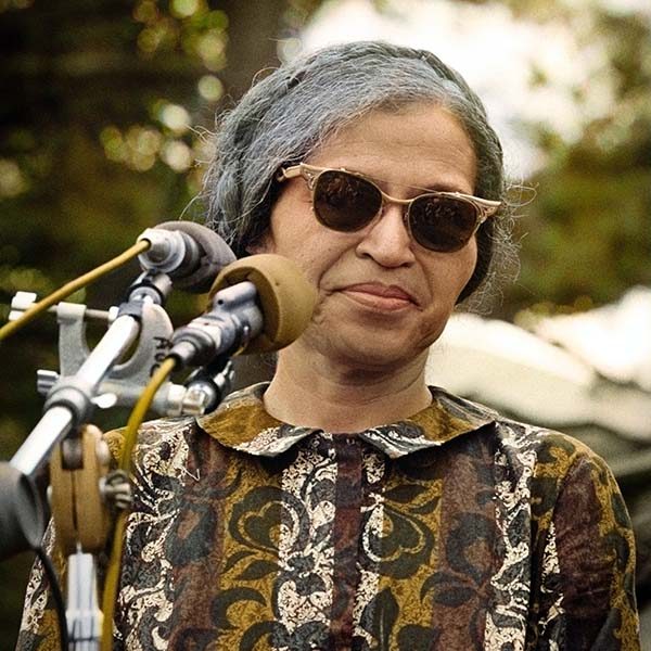 Rosa Parks standing outside in front of a microphone. She wears sunglasses and a paisley print dress