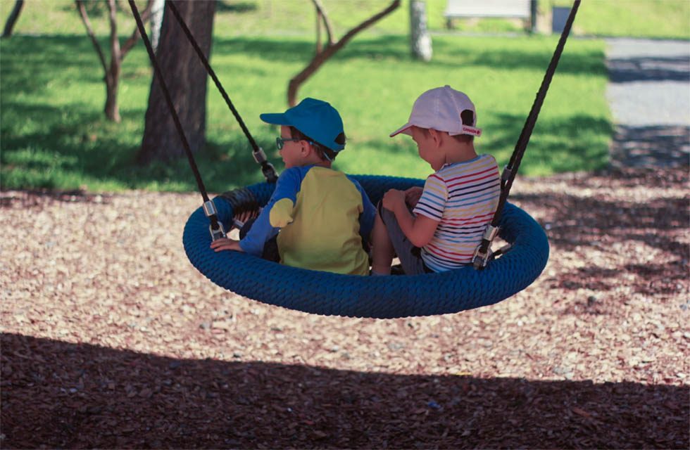 Two preschool-aged boys in brightly-colored t-shirts and baseball hats sit in a tire-style tree swing on a sunny day.