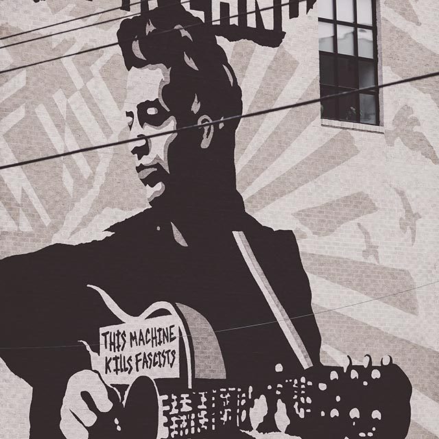 A black and white mural of a young Woody Guthrie playing guitar with a sign on guitar saying, 